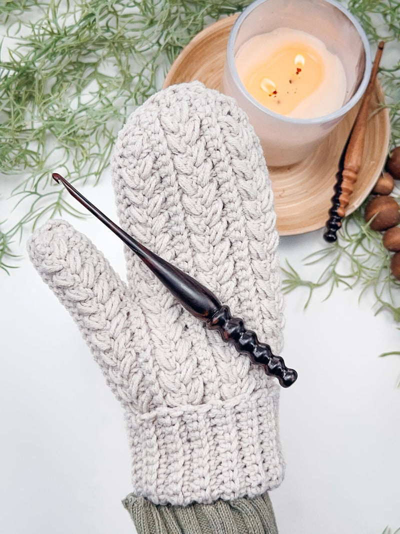 Light gray crochet mitten with braided texture worn on a hand, with a wooden crochet hook resting on top