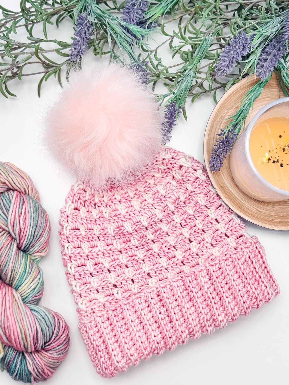 A pink crochet beanie with a fluffy pom-pom, surrounded by colorful yarn and lavender sprigs, rests beside a wooden tray holding a cup of cappuccino—an ideal handmade gift for someone who appreciates cozy aesthetics.
