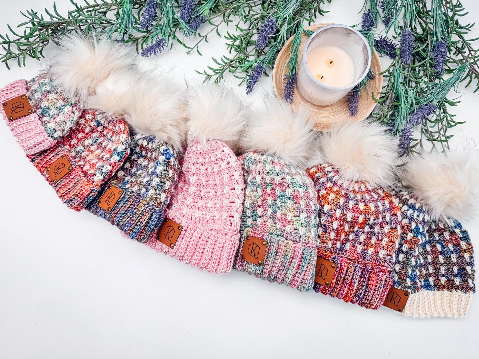 Seven colorful crocheted hats with pom-poms are arranged in a row, each boasting leather patches. They sit beneath sprigs of lavender and a lit candle, making for the perfect handmade gift display.