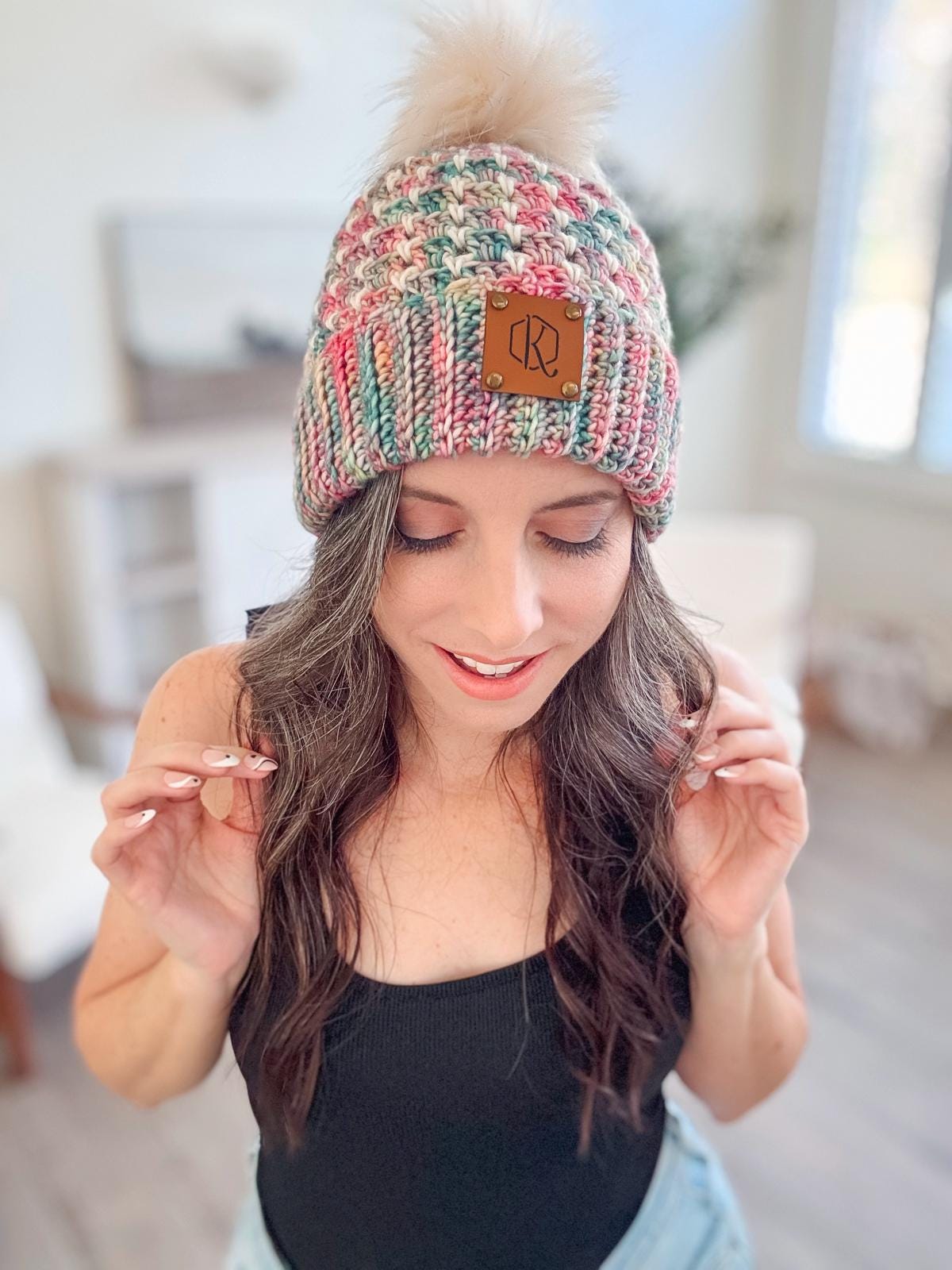 A woman with long brown hair wearing a multicolored crochet hat with a pom-pom. She is indoors, dressed in a black top, and holding the hat's brim with both hands. Her style is accentuated by vintage clothes that add a timeless charm to her ensemble.