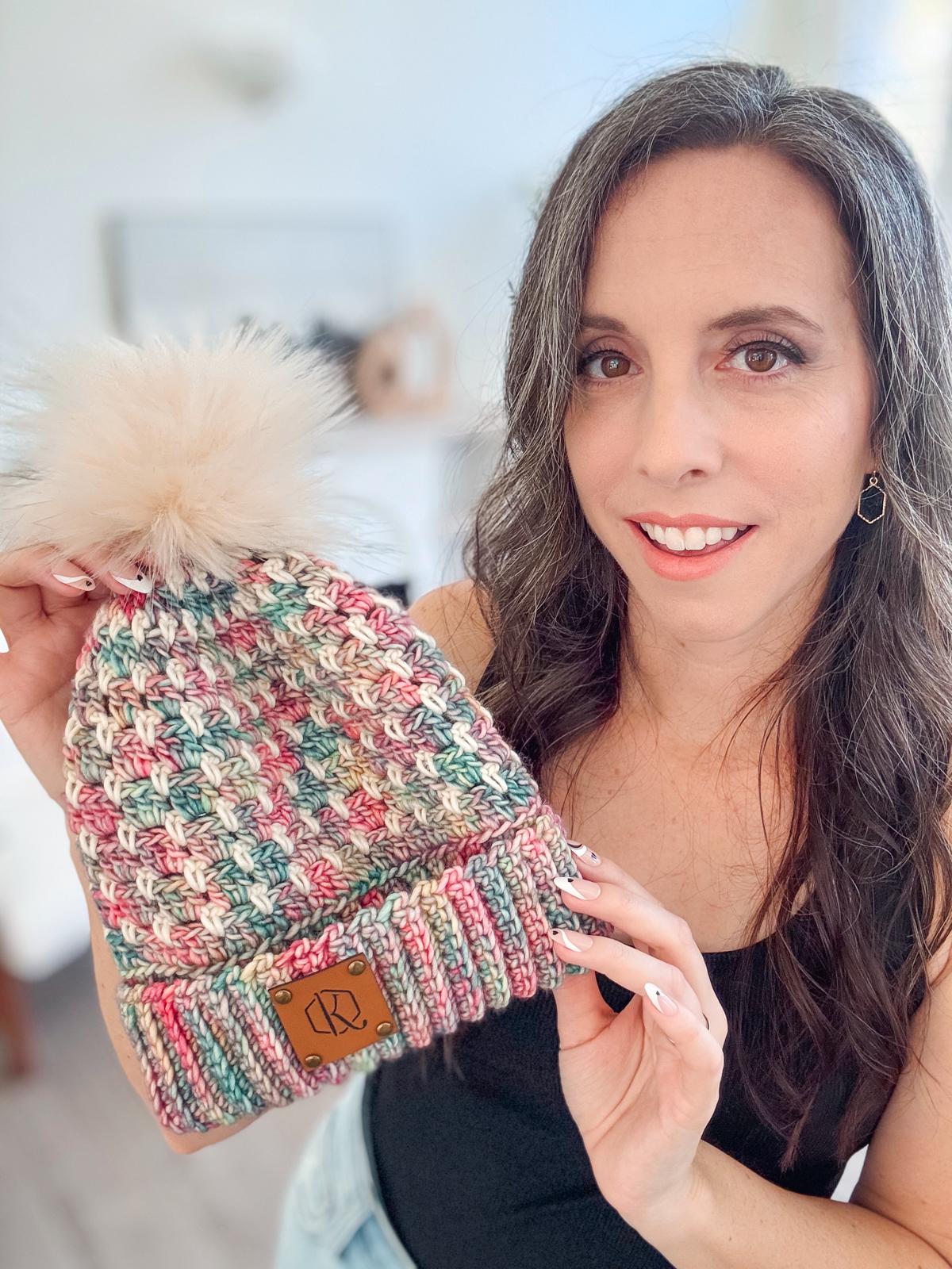 In a well-lit room, a woman smiles while holding a multicolored crocheted beanie with a fluffy pom-pom, reminiscent of vintage clothes.