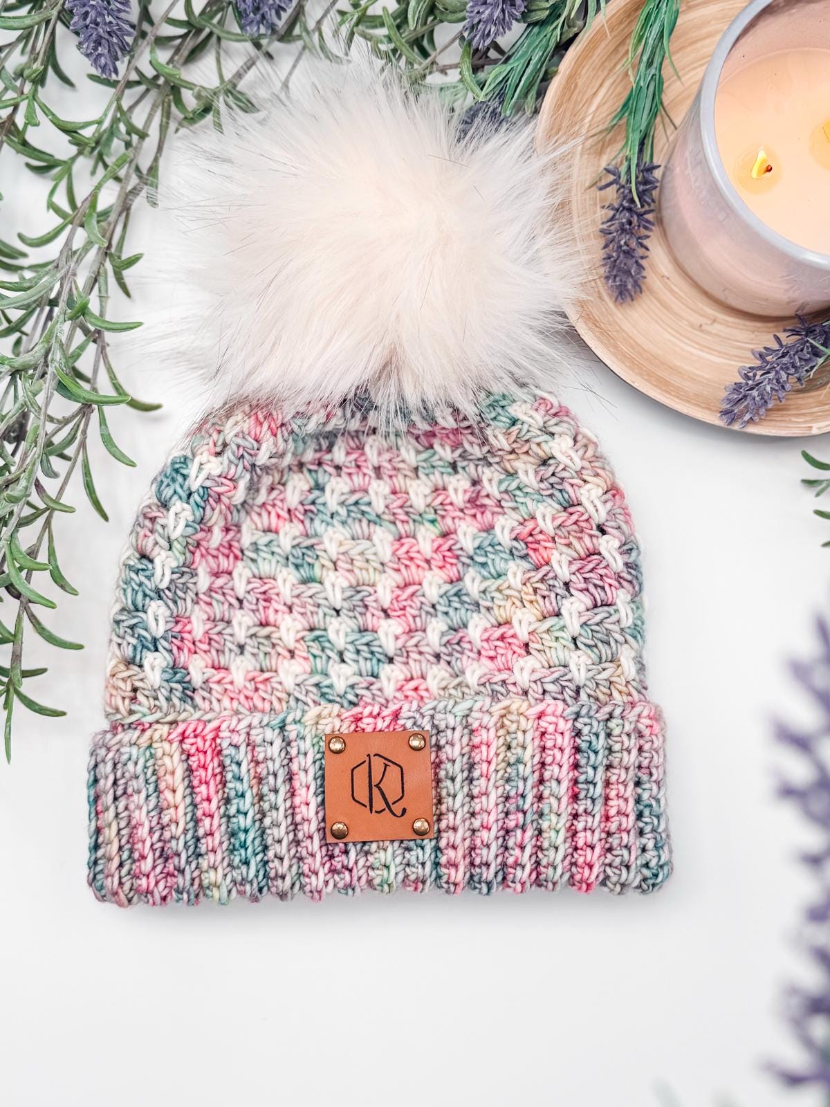 A multicolored crochet hat with a white pom-pom and a leather patch sits on a surface, surrounded by lavender sprigs, a lit candle.