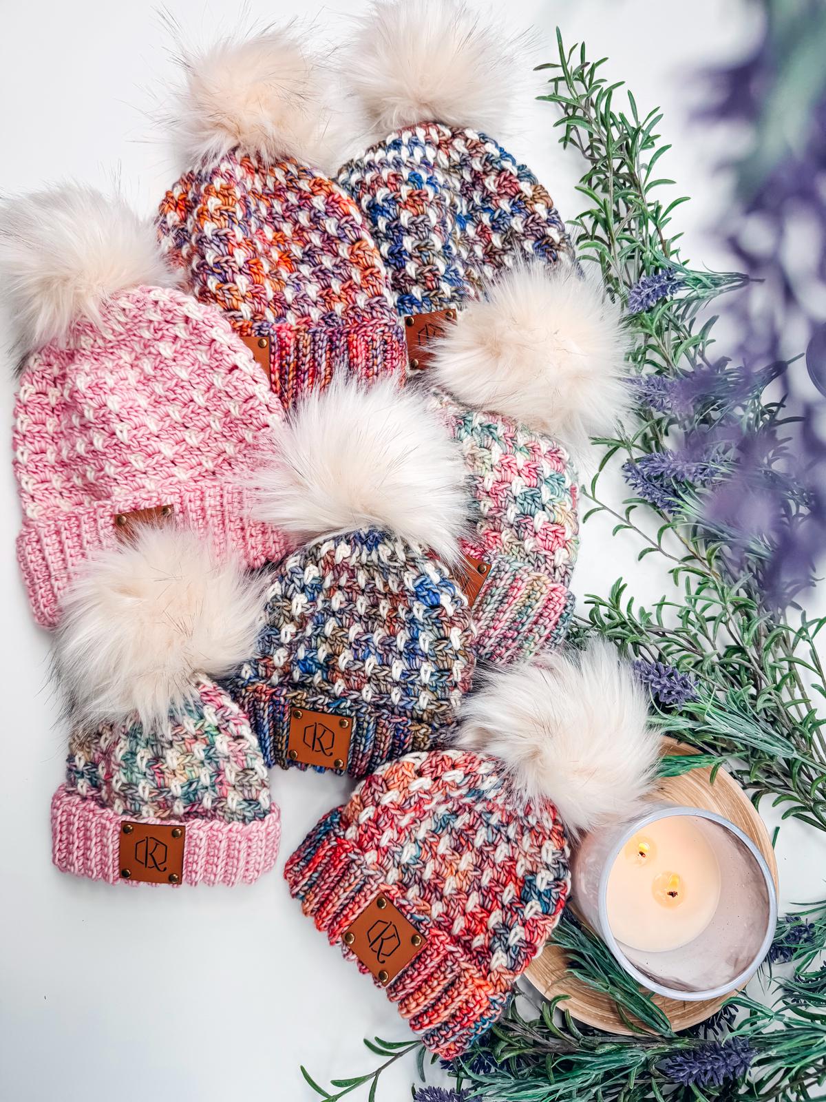 Six colorful crochet hats with pom-poms are arranged on a surface. Sprigs of greenery and a lit candle set the scene, making it a perfect handmade gift.