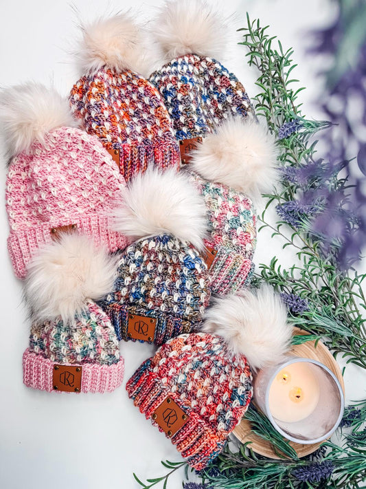 Six colorful crochet hats with pom-poms are arranged on a surface. Sprigs of greenery and a lit candle set the scene, making it a perfect handmade gift.
