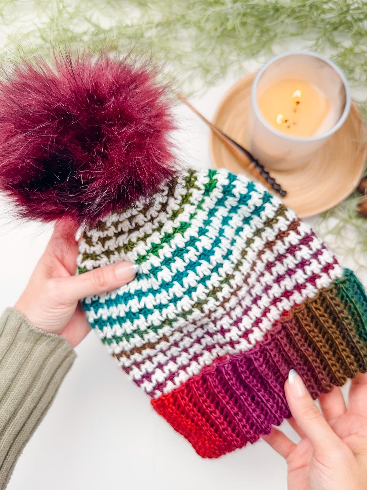 A person holds a colorful, knitted hat with a large maroon pom-pom, perfect as a handmade gift. A lit candle and a green plant are in the background.