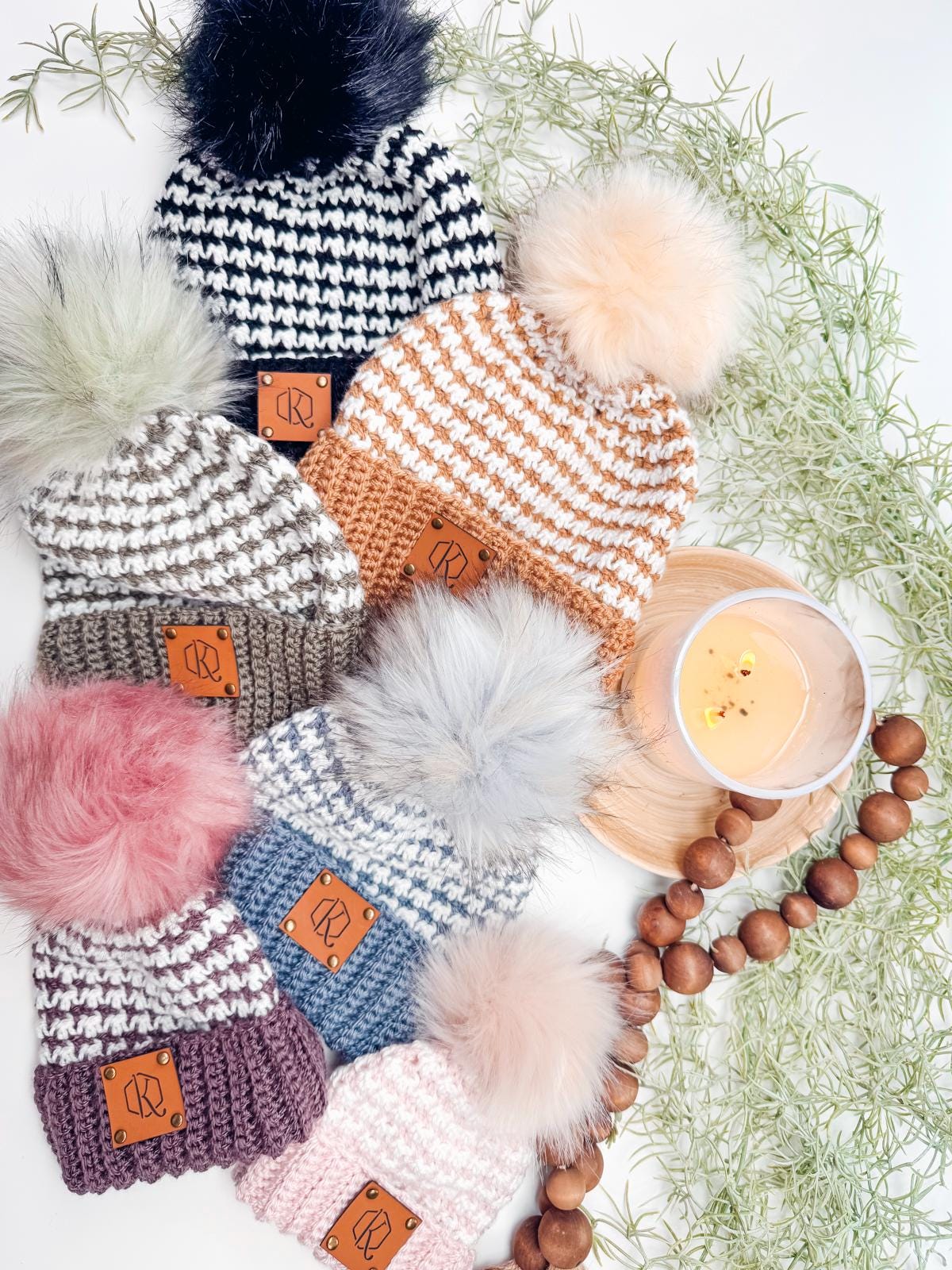 A collection of six knitted pom-pom hats in various colors, arranged with a candle and wooden beads on a white background, adorned with greenery. Perfect as a handmade gift, this charming display captures the warmth of cozy elegance.