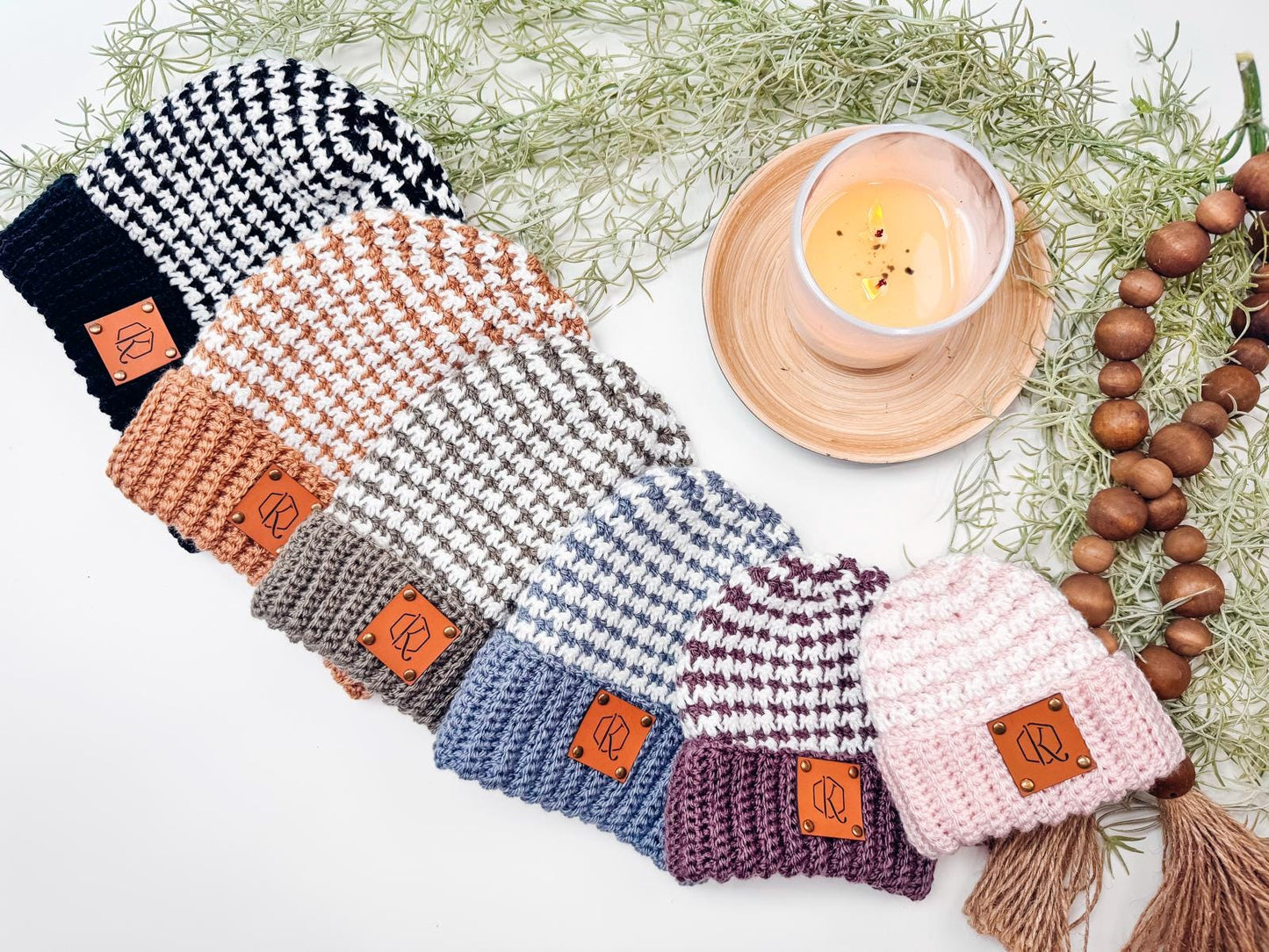 Six knitted beanies in various colors are displayed in a row next to a lit candle and decorative greenery on a white surface, offering the perfect setting for selecting a handmade gift.