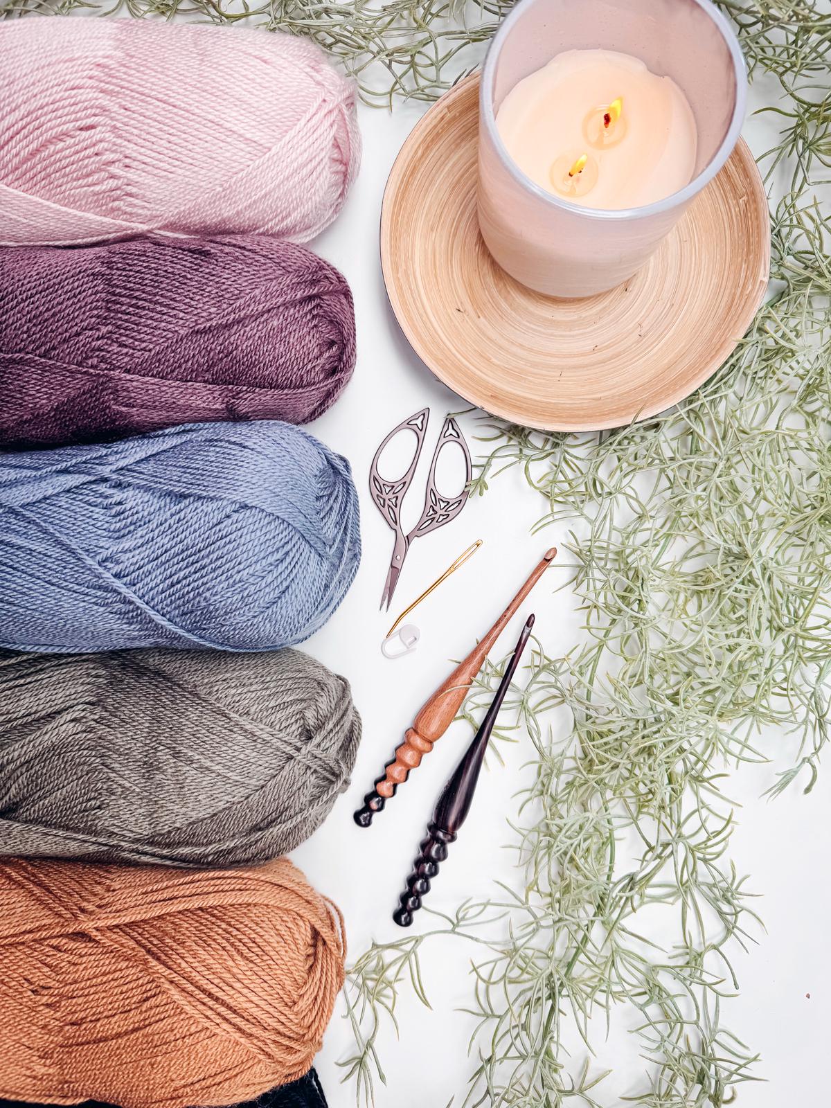 Four skeins of yarn, scissors, two crochet hooks, and a candle are artfully arranged on a white surface, surrounded by lush greenery. Perfect for crafting a handmade gift that reflects your personal touch and creativity.