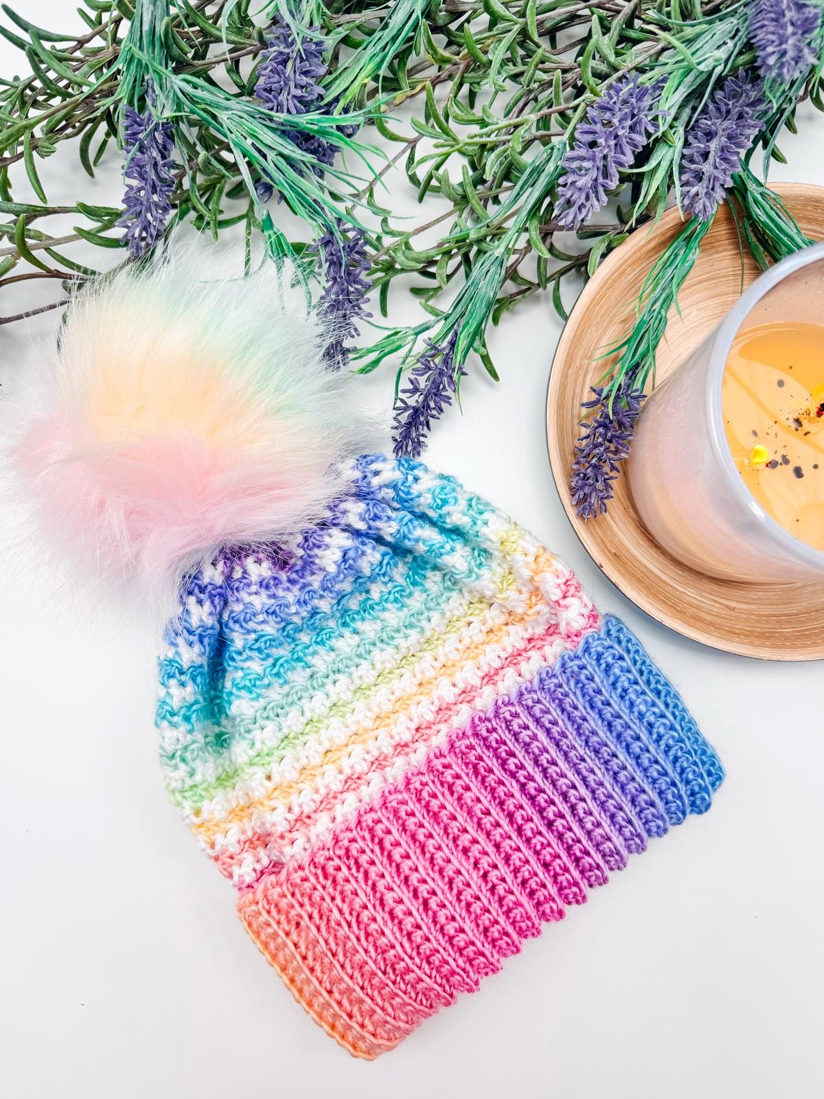 A colorful knitted beanie with a fluffy pom-pom, perfect as a handmade gift, sits on a table next to a bowl and sprigs of lavender.