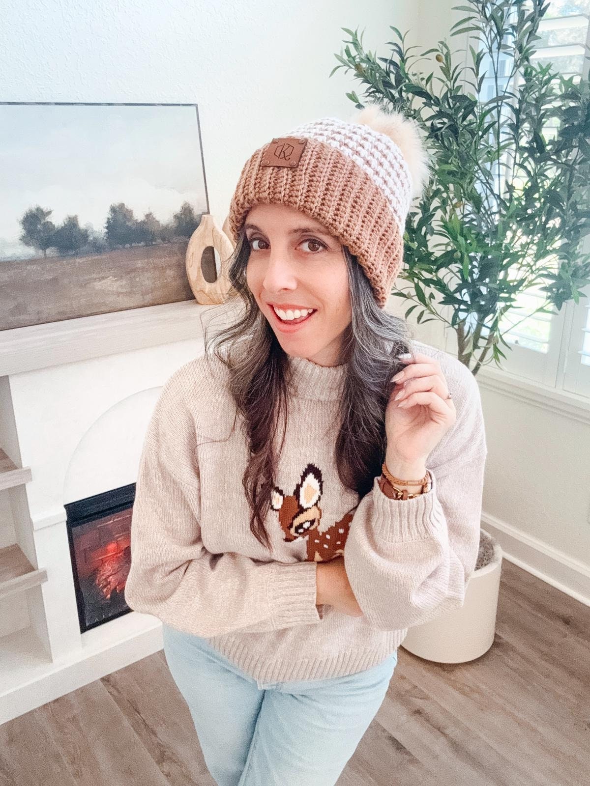 A woman in vintage clothes, wearing a brown knit hat and a sweater with a deer design, poses indoors near a fireplace and a potted plant.