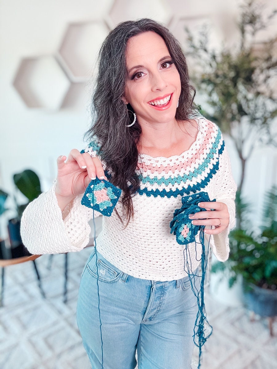 A woman in a white and teal knit sweater shows a crocheted square, holding more squares and yarn in her other hand, standing in a cozy room with plants and geometric wall decor.
