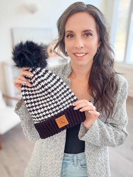A person stands indoors, holding a black and white knitted beanie with a pom-pom, perfect as a handmade gift. The beanie features a brown patch, complementing the gray textured jacket and jeans worn by the stylish individual.