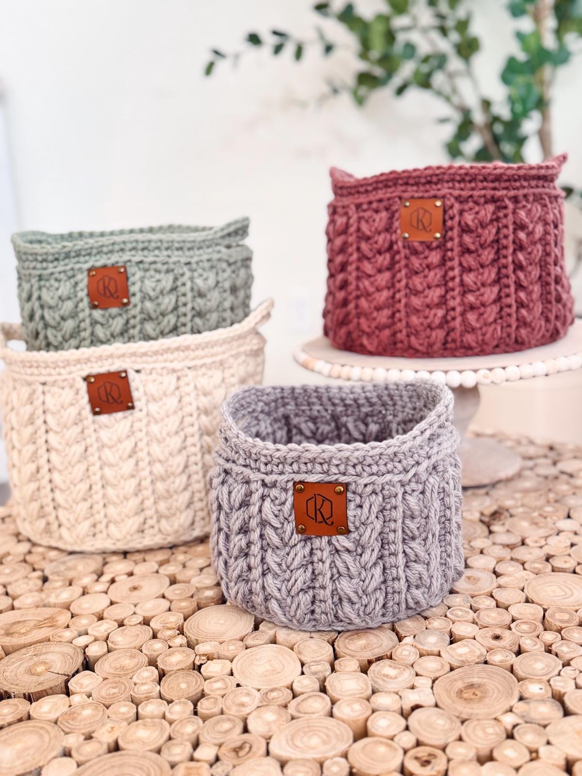 Four crochet baskets in green, pink, cream, and gray rest on a wooden table. Each basket, perfect as a handmade gift, features a leather patch with a circular logo.
