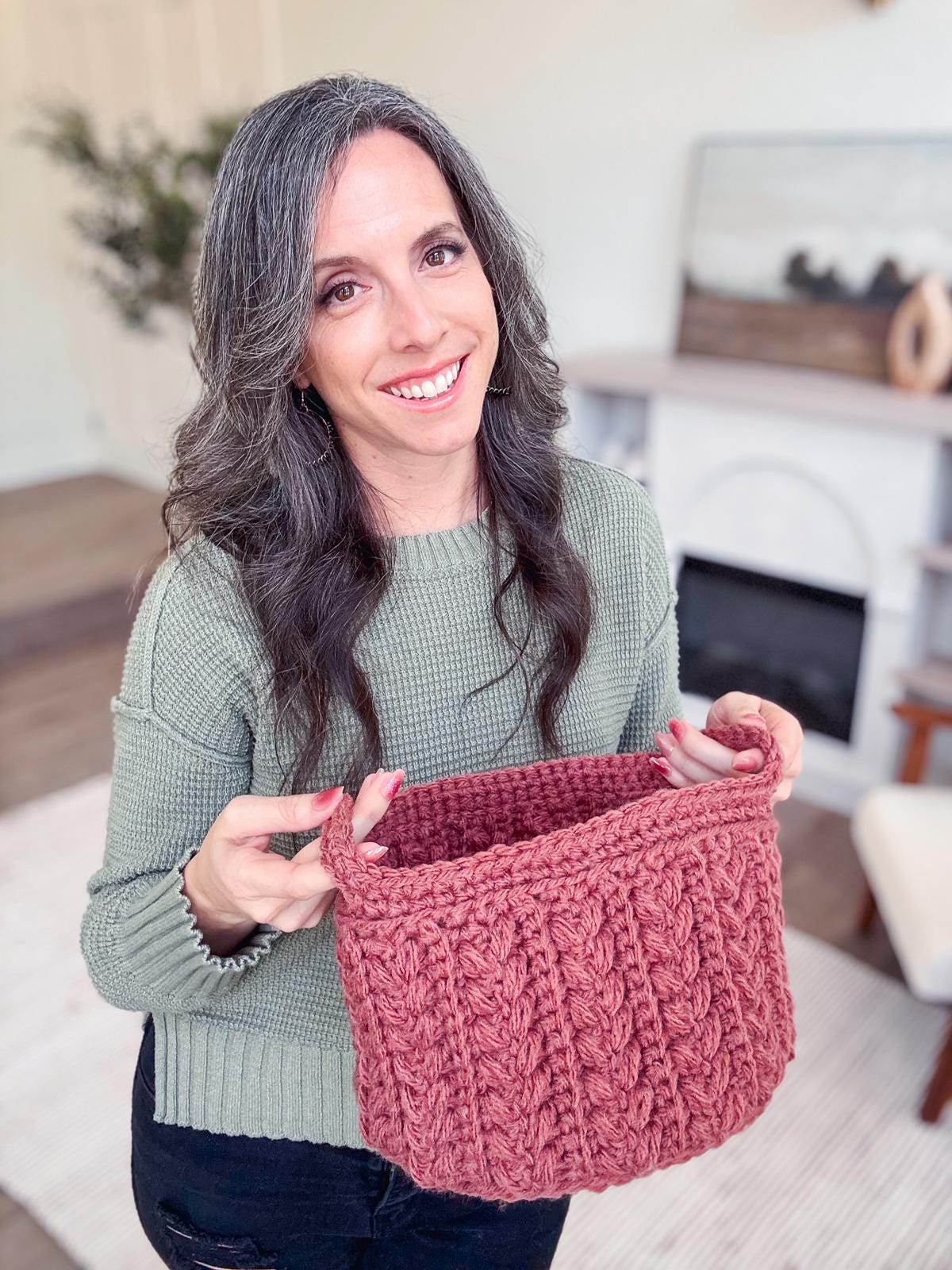 A person with long hair and a green sweater holds a pink crochet basket, standing indoors with a fireplace and decorative background, showcasing their style that blends vintage clothes with the charm of unique jewellery.