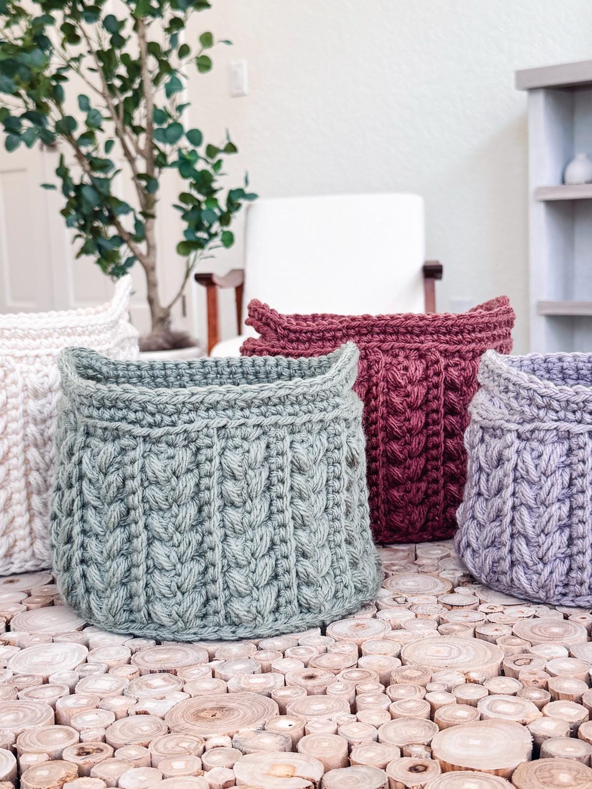 Three crocheted baskets in green, red, and purple sit on a wooden surface, with a plant and white chair in the background. These baskets make an ideal handmade gift for those appreciating artisanal charm.