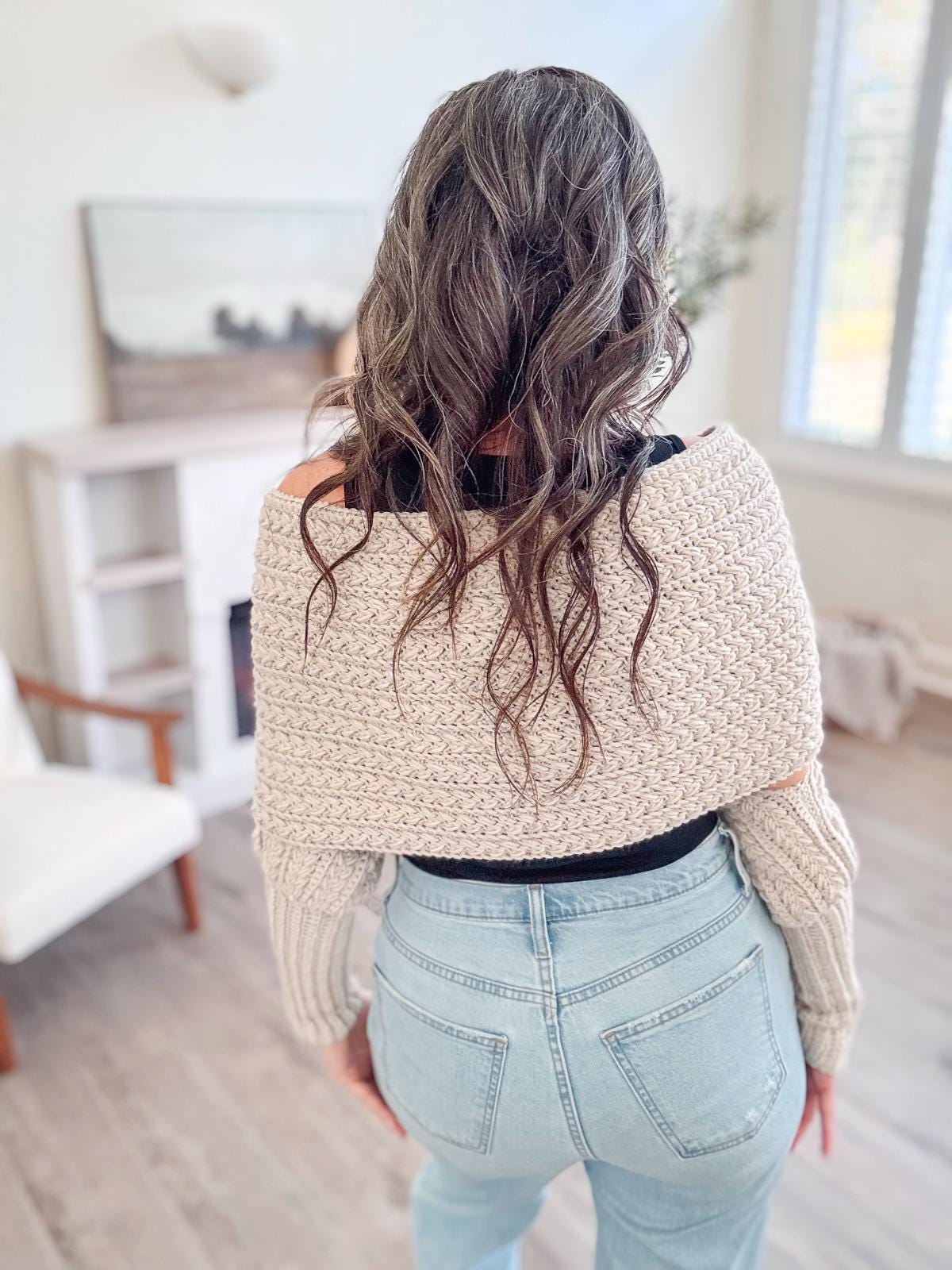 A person with wavy brown hair in a beige crochet sweater, adorned with unique jewellery, stands in blue jeans in a room featuring a chair and an intriguing painting.