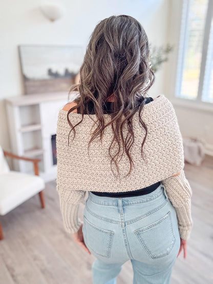 Back view of a cream crochet sweater scarf worn off the shoulders by a woman with long wavy hair and jeans