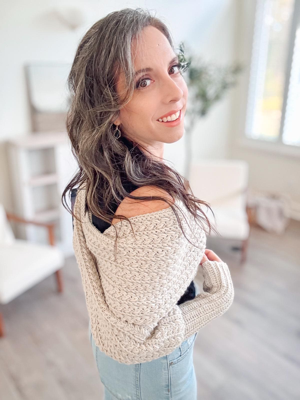 A woman with long hair and a light crochet sweater looks over her shoulder, smiling, in a bright room adorned with vintage clothes and a plant, exuding warmth and charm.