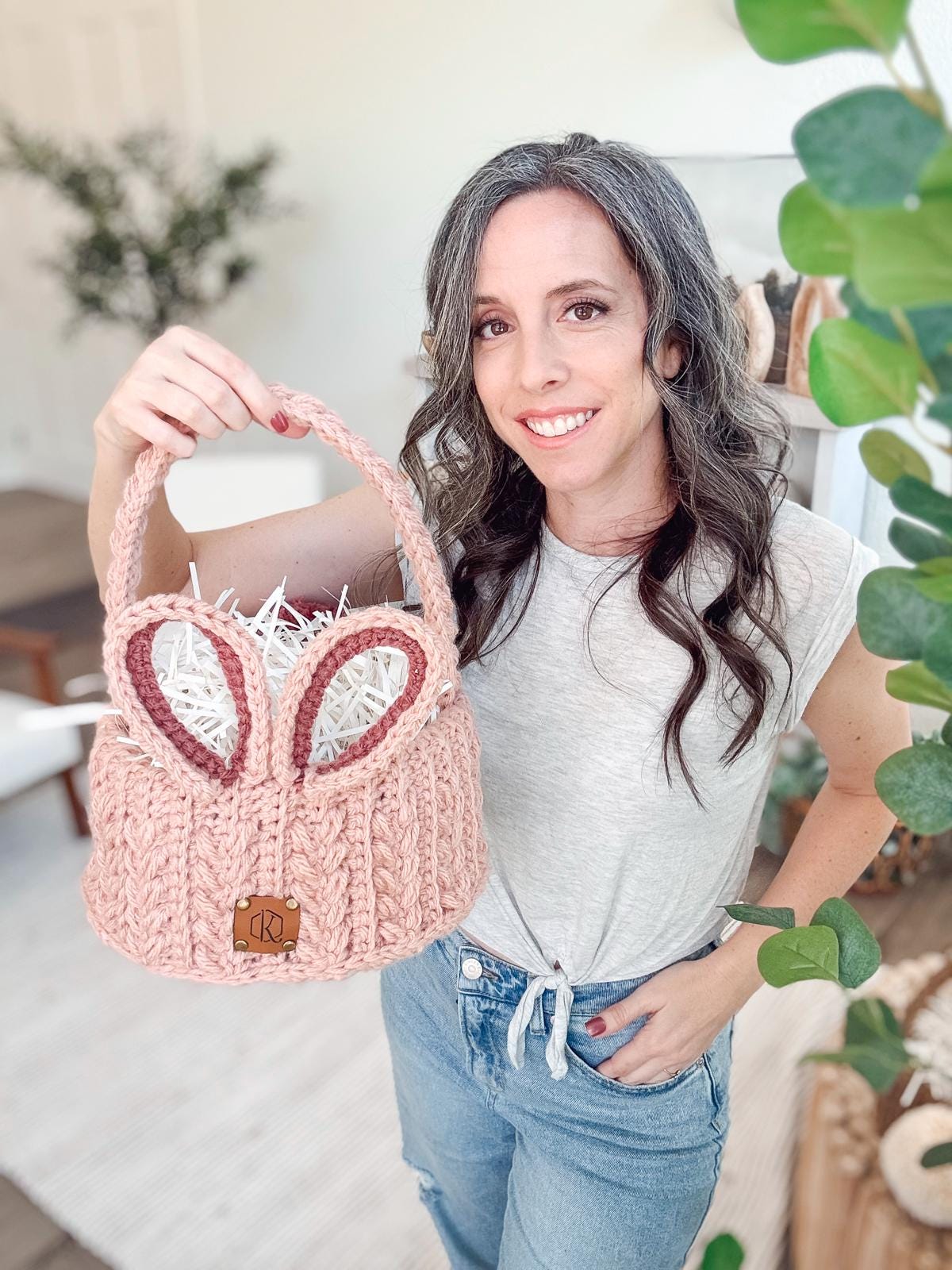 A person holding a crochet pink basket with bunny ears, standing in a room adorned with lush plants —a charming tableau of handmade Easter basekt crochet gifts.