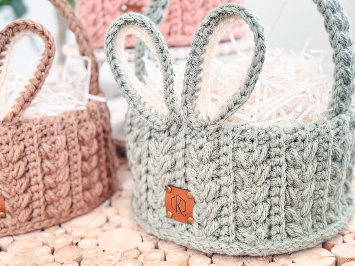 Crocheted baskets with handles, one in green resembling bunny ears, are filled with shredded paper on a wooden surface. These charming creations make for a delightful handmade gift, adding a touch of whimsy and creativity to any setting.