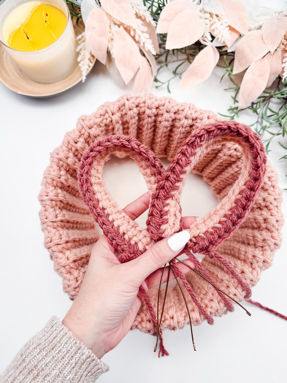A hand holding pink bunny ears above a coiled crocheted piece, near a lit candle and decorative leaves, showcases the beauty of handmade gifts.