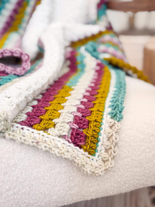 Close-up of a crocheted blanket draped over a chair, an ideal handmade gift. The blanket features stripes of white, mustard yellow, teal, pink, and purple with a textured pattern.