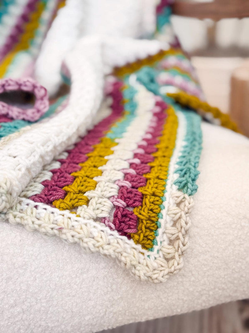 Block Stitch Blanket crochet pattern: close-up of a cream crochet blanket edge with pink, mustard, teal, and white block stitch stripes on a white chair