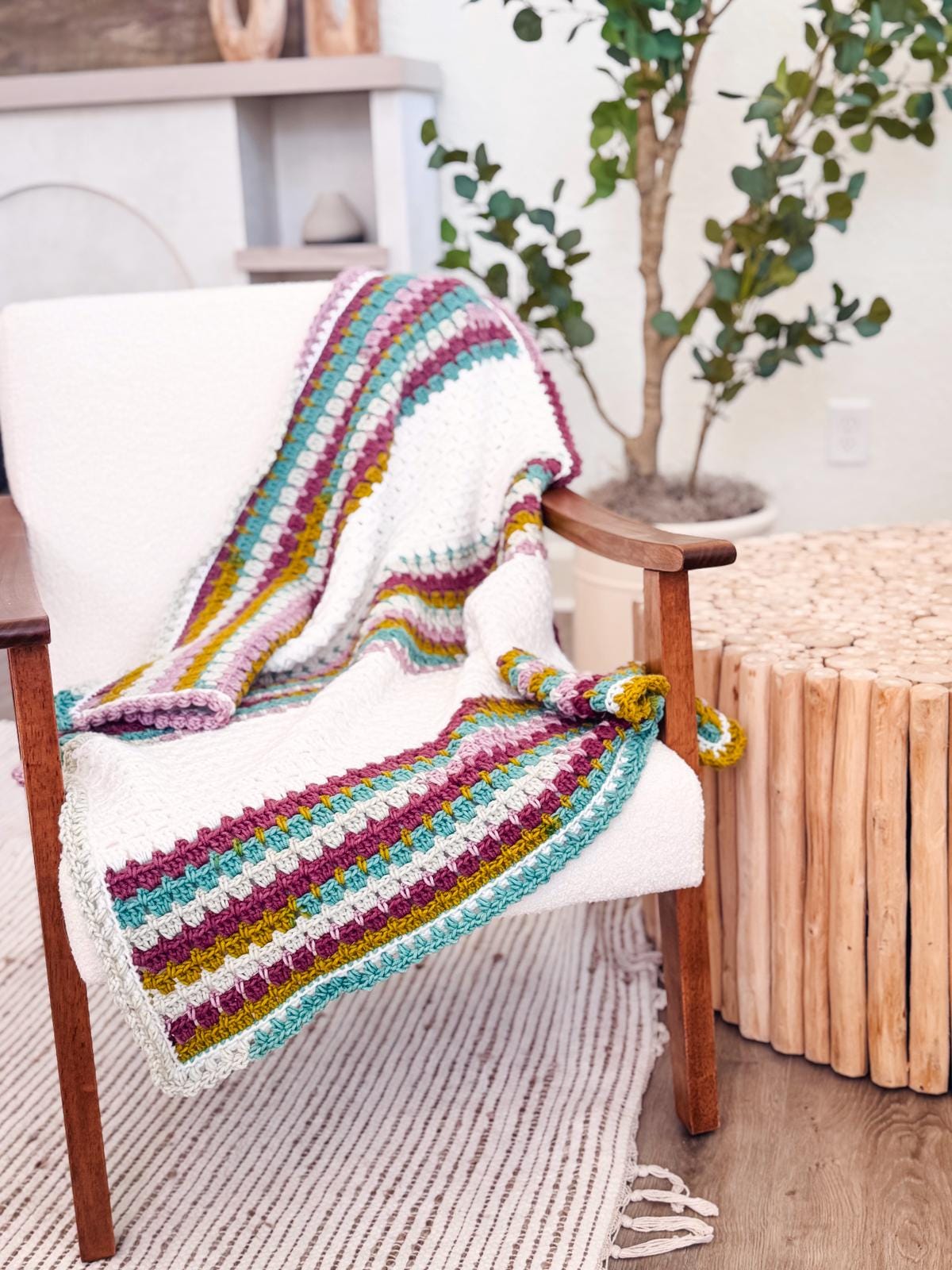 A multicolored crochet blanket draped over a white armchair next to a wooden table, offering the perfect touch of charm for your home. A plant stands in the background on a striped rug, reminiscent of an inviting space filled with handmade gifts and unique jewelry treasures.