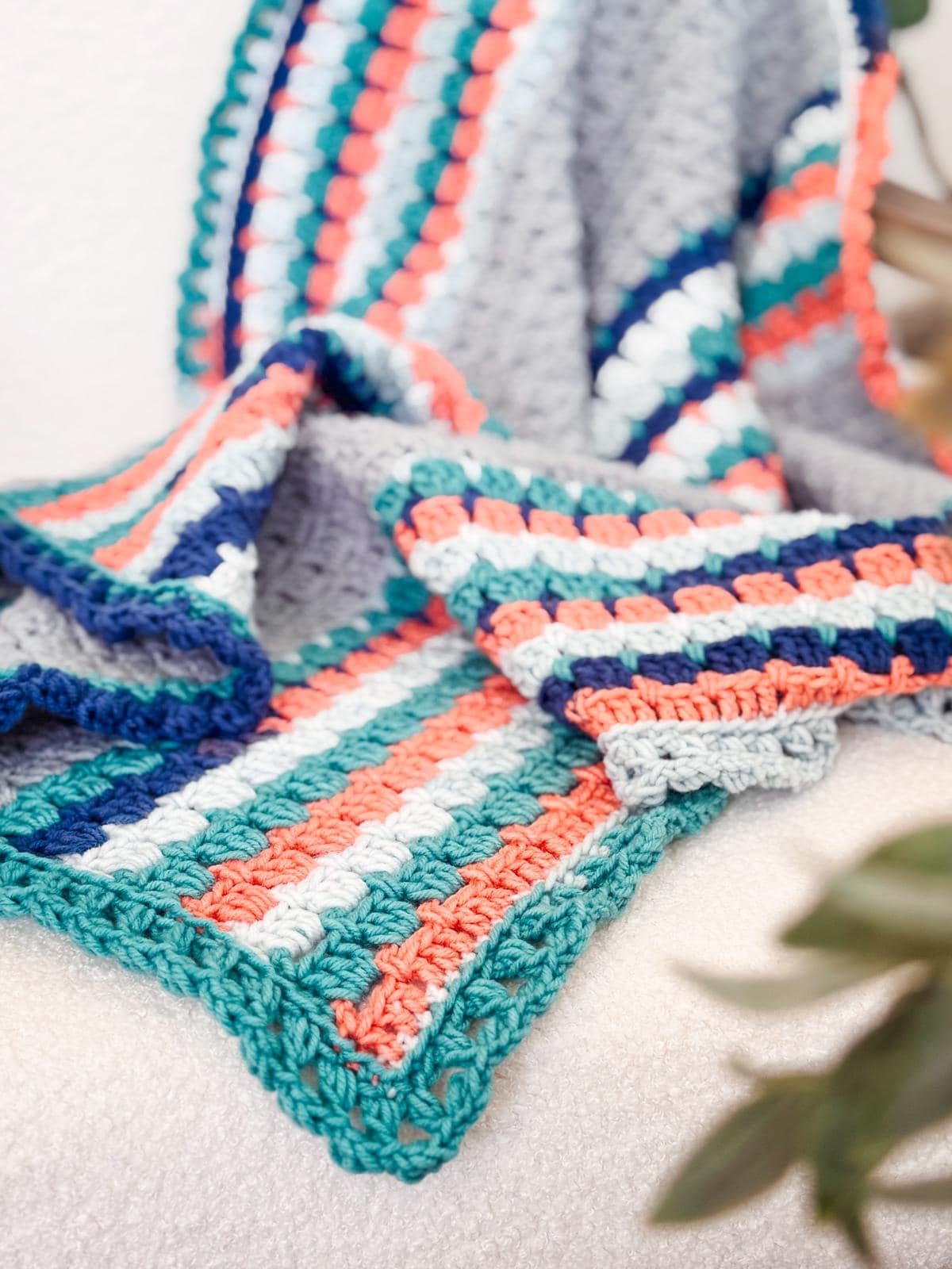 A colorful striped crochet blanket, like a vintage handmade gift, is draped over a light-colored surface with green leaves in the foreground.