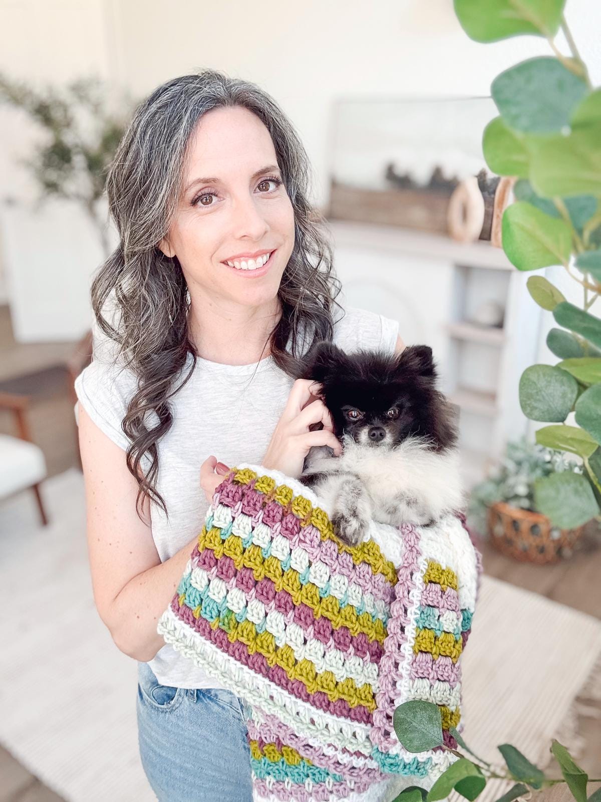 A woman wearing jeans cradles a small black and white dog in a multicolored crochet blanket. They stand together in a bright room adorned with plants, home decor, and unique jewelry pieces.