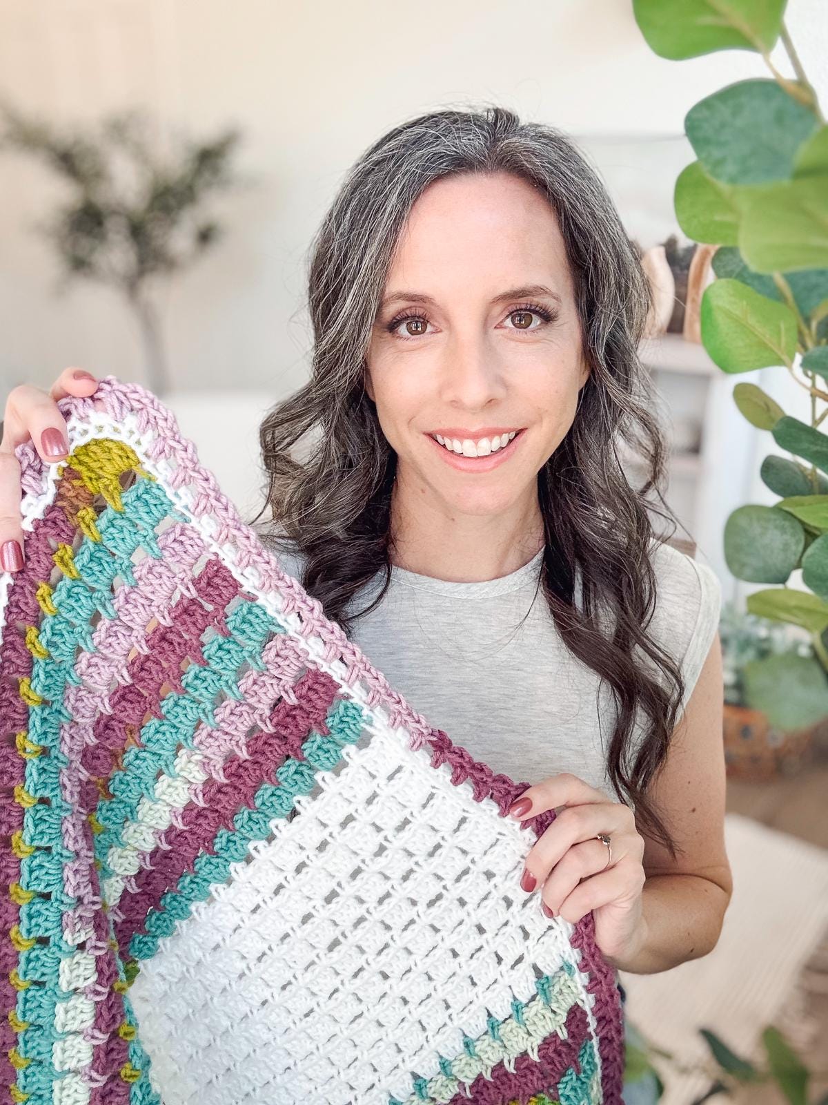 A person stands indoors, cradling a multicolored crocheted blanket. Lush plants adorn the background, creating a serene scene that highlights the charm of this handmade gift.
