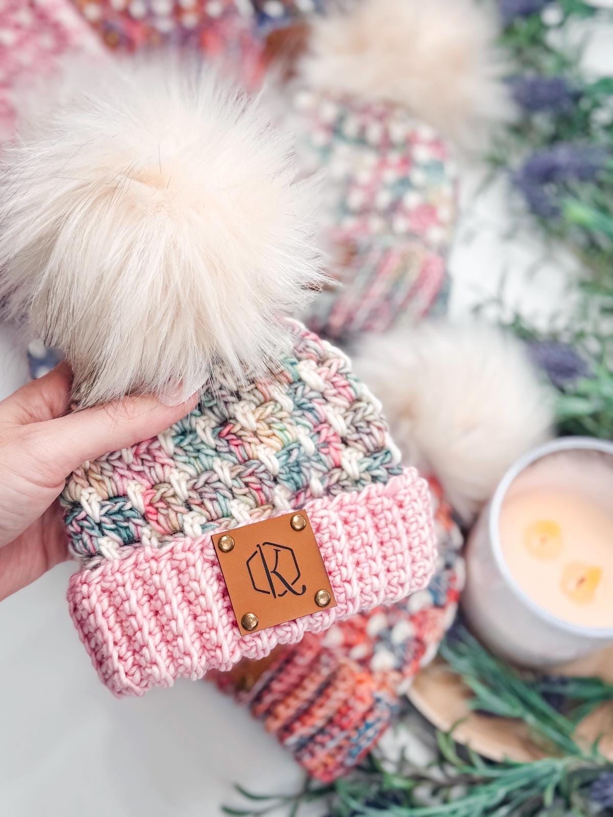 A hand holds a crocheted beanie with a pom-pom and leather patch, perfect as a handmade gift. In the background, other beanies accompany dried flowers and a lit candle, creating a cozy scene.