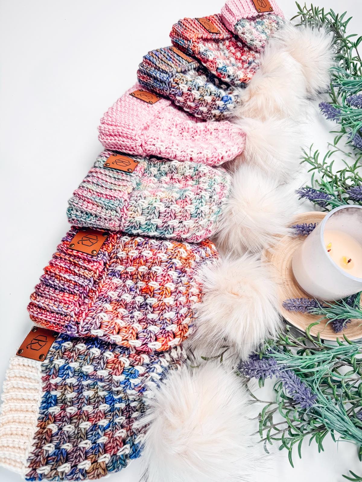 A selection of colorful crocheted hats with faux fur pom-poms, beautifully arranged next to a lit candle and sprigs of greenery, makes for the perfect handmade gift.