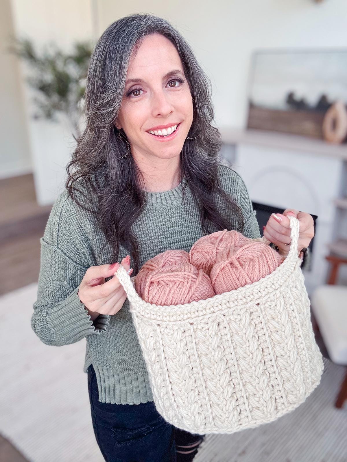 A woman with long hair holds a crochet nesting basket containing several skeins of yarn in a cozy room with a fireplace, each skein destined to become a handmade gift.