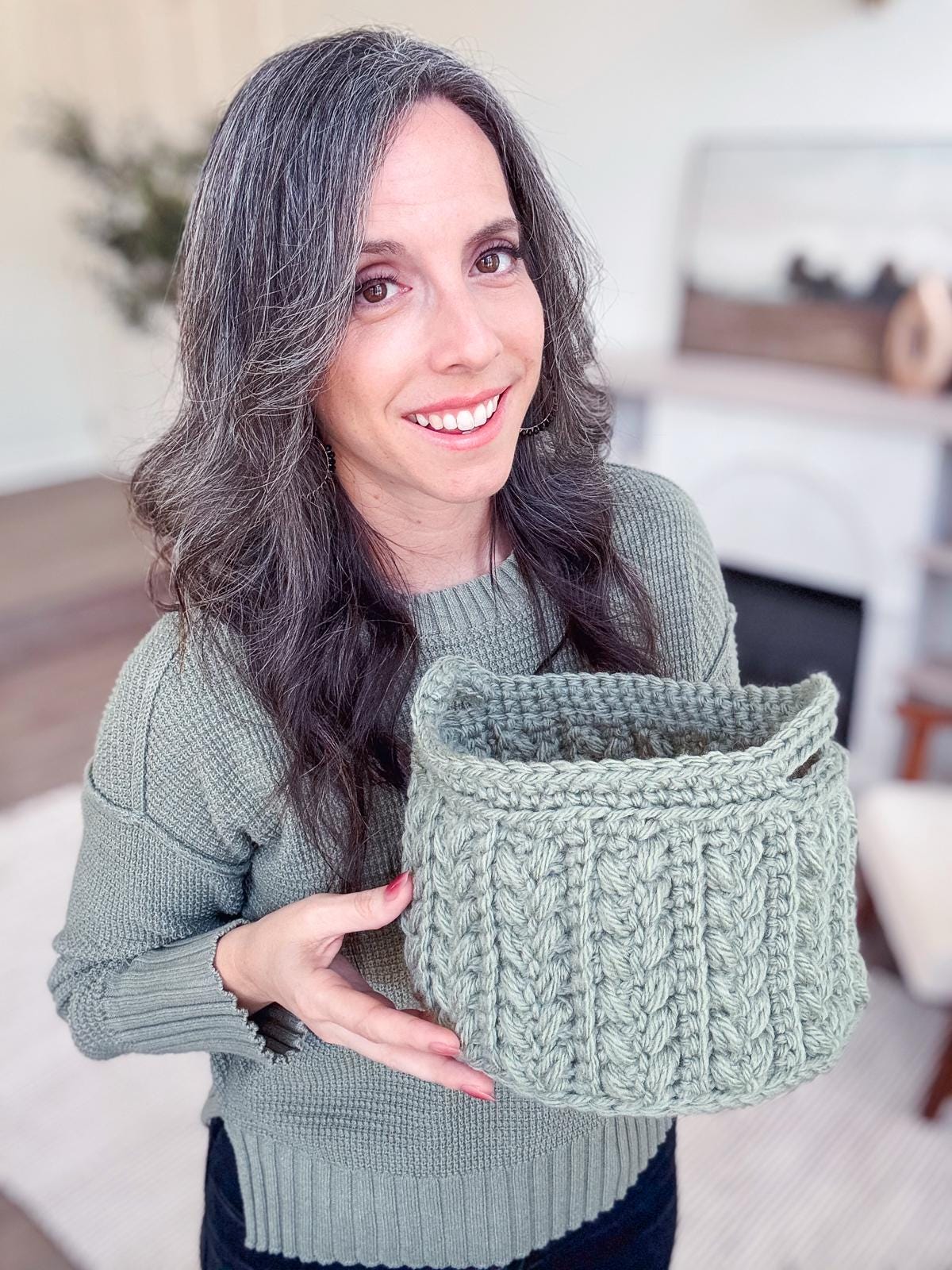 A person in a green sweater holds a beautifully crocheted, light-green basket with a textured pattern, smiling at the camera. This handmade gift perfectly complements the blurred interior setting in the background.