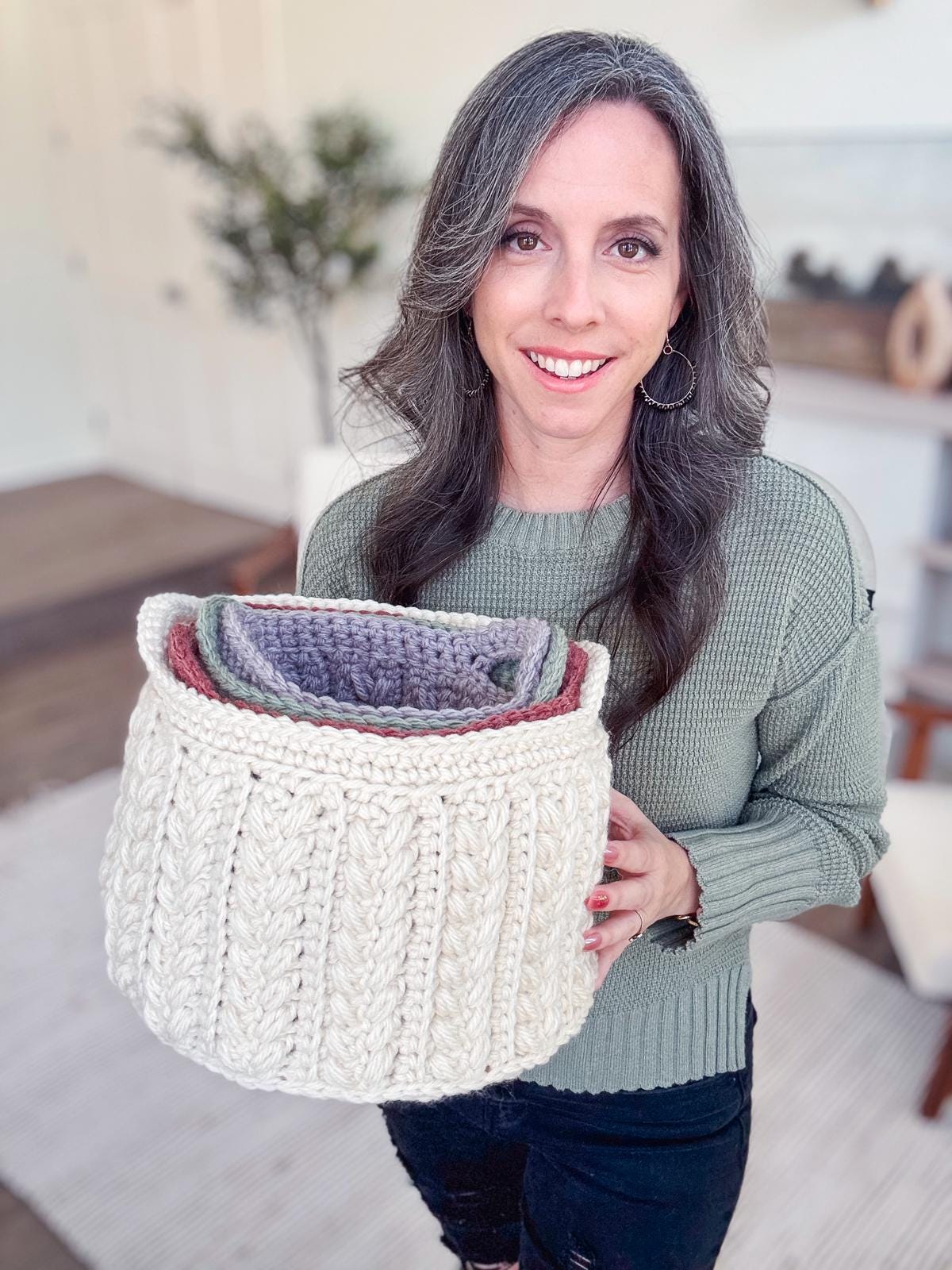 A woman with long hair holds a large, cream-colored crocheted basket. She wears a green sweater and stands in a room adorned with unique jewelry and furniture, alongside a plant, creating the perfect setting for displaying handmade gifts. Crochet pattern