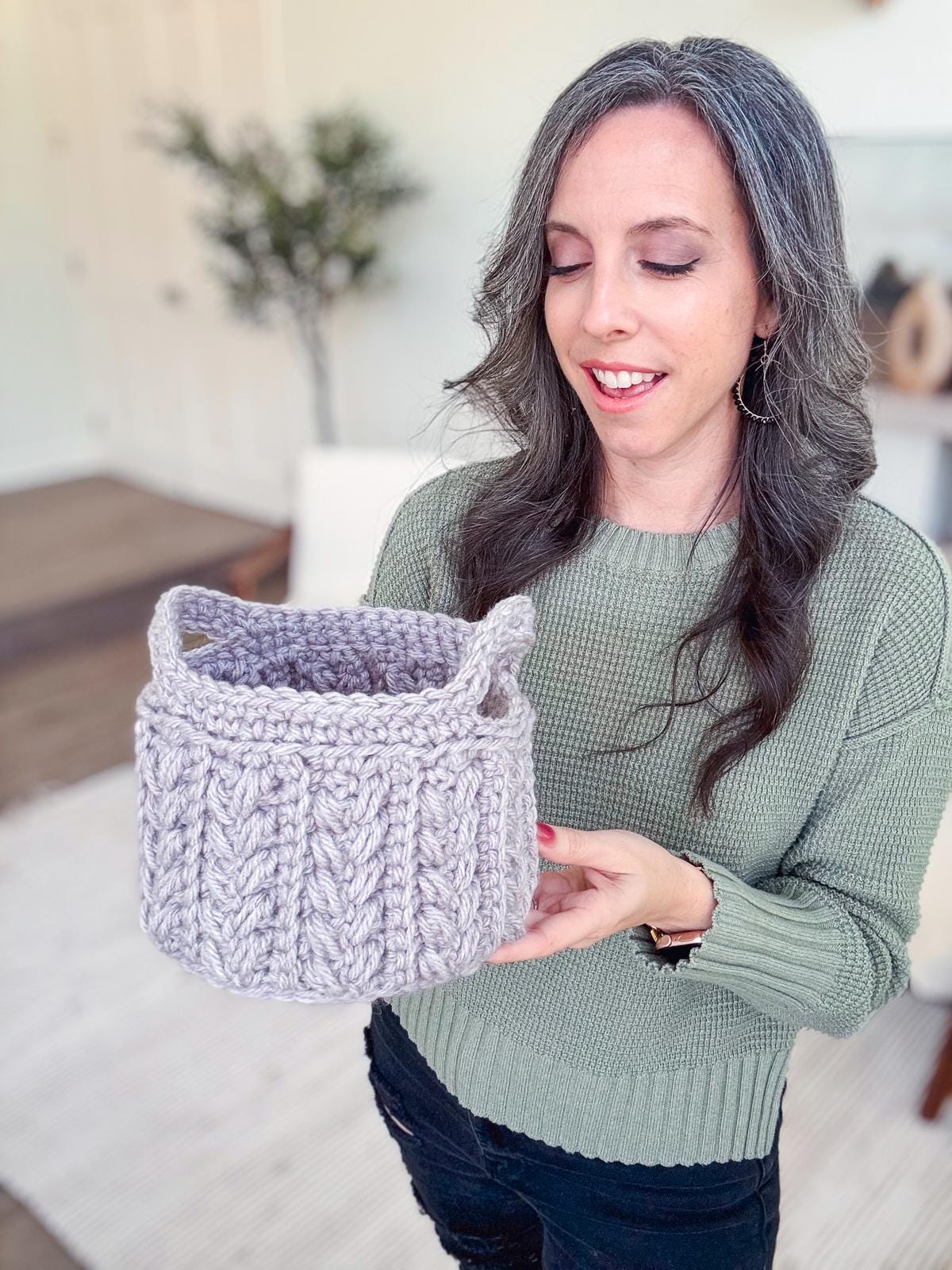 A person with long dark hair, in a green sweater adorned with unique jewellery, holds a gray crochet basket with handles in a neutral-toned room.