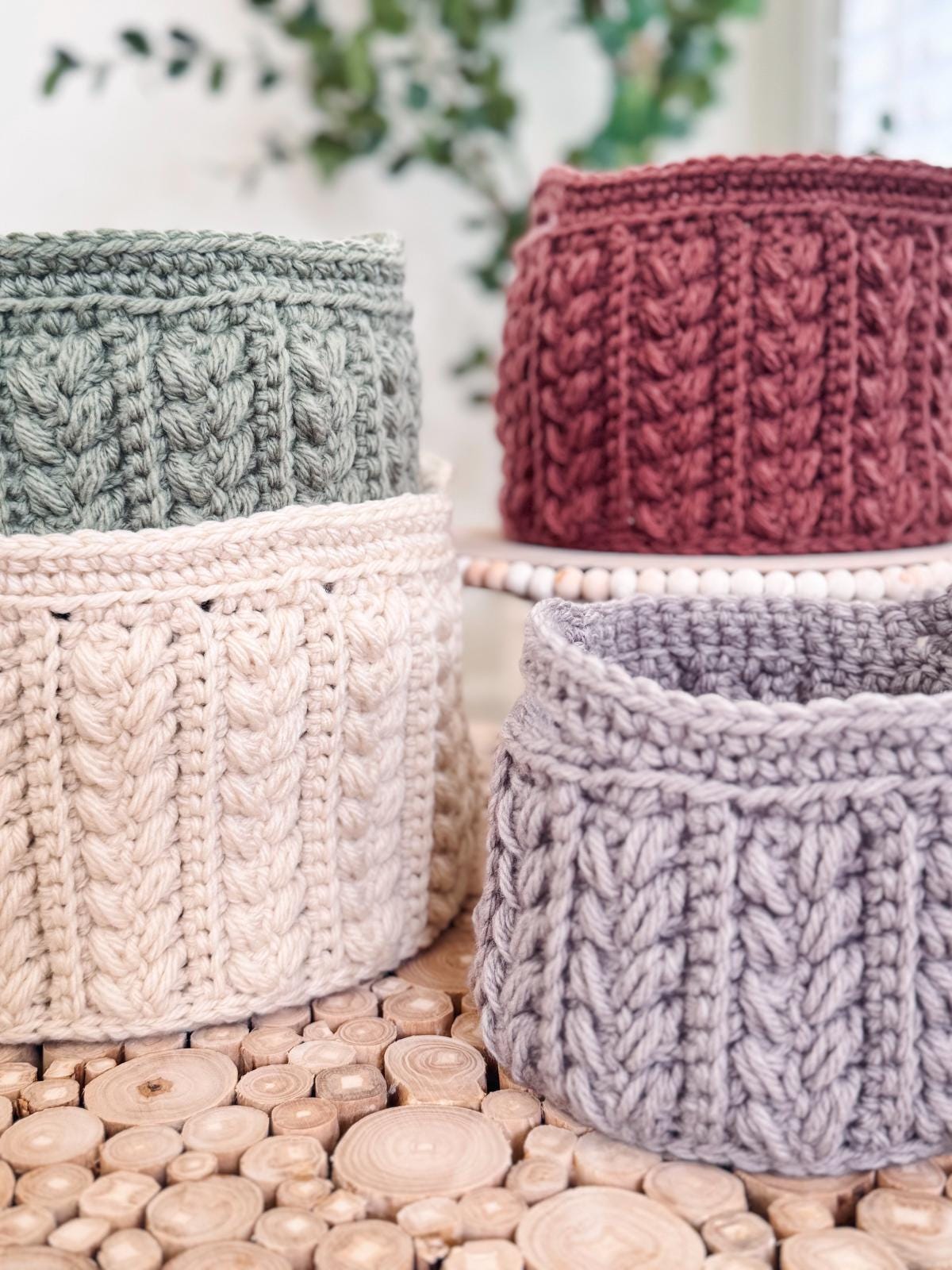 Four crocheted nesting baskets in green, red, beige, and gray sit on a wooden surface, with plants in the background. These charming baskets make an ideal handmade gift for any occasion. Crochet Pattern