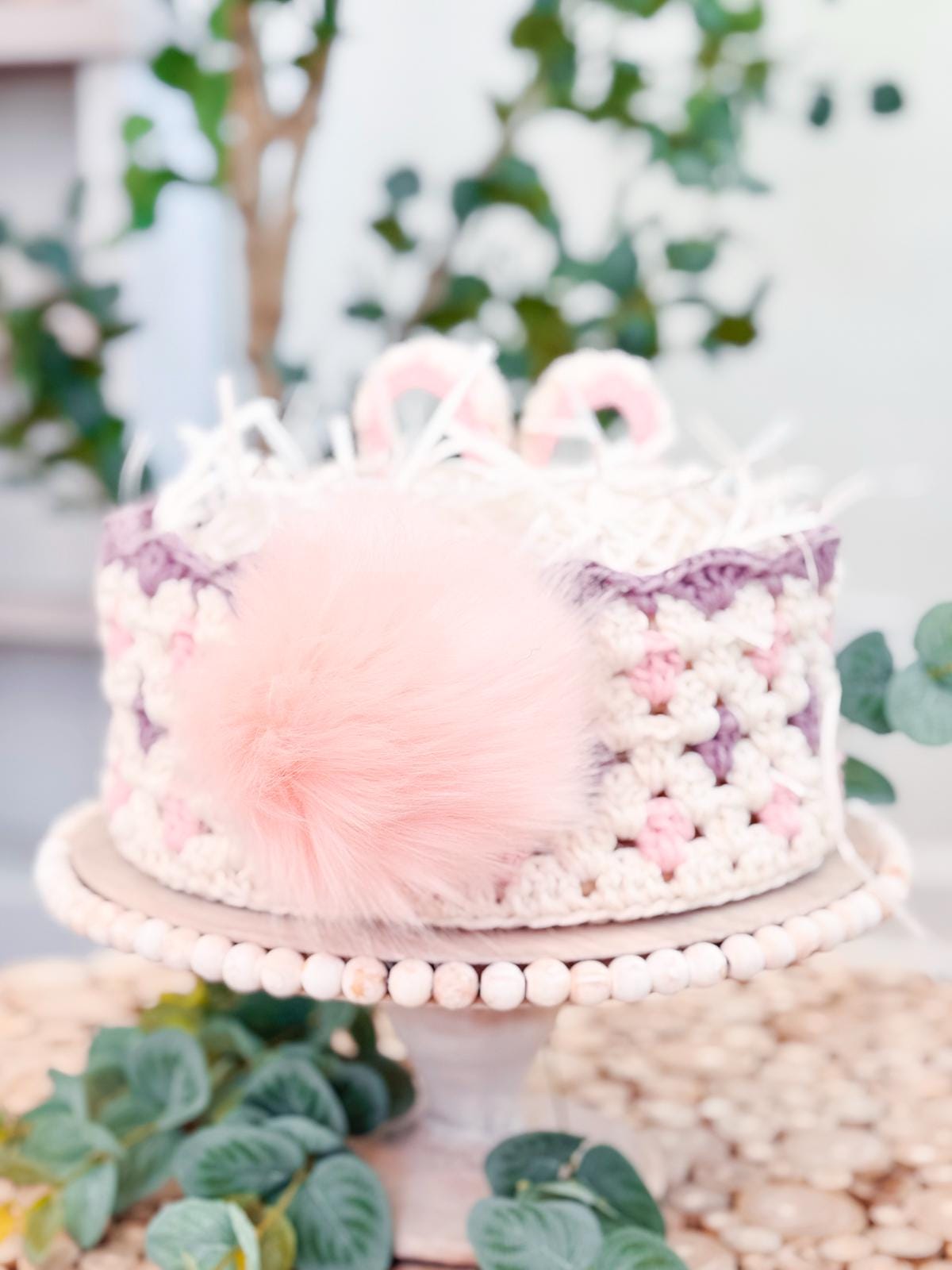 A crocheted hat with a pink pom-pom is displayed on a wooden stand, making the perfect handmade gift. It's surrounded by lush greenery, adding a touch of nature's charm.