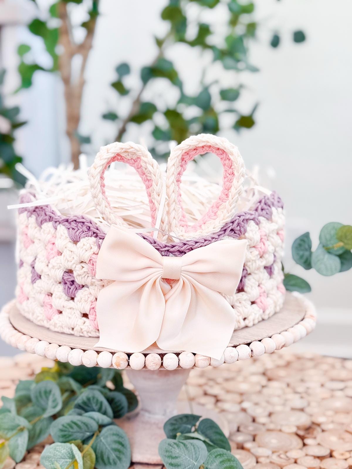 A crocheted basket with bunny ear handles and a large pink bow is set on a wooden tray, surrounded by greenery, making it the perfect handmade gift.