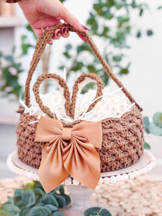 A hand holds a crocheted basket adorned with a large brown bow, filled with white shredded paper, set on a round platter amidst lush greenery. This charming scene evokes the simplicity and charm of handmade gifts for Easter.