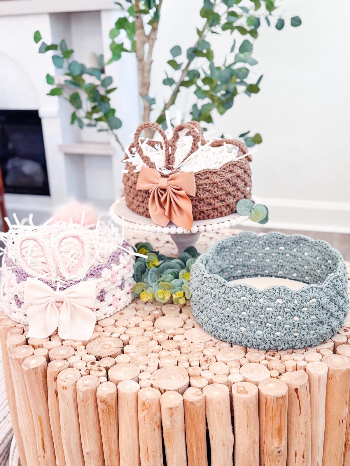 Three woven crochet easter bunny baskets with bows and decorative fillings sit on a wooden table, surrounded by greenery, each resembling a handmade gift.