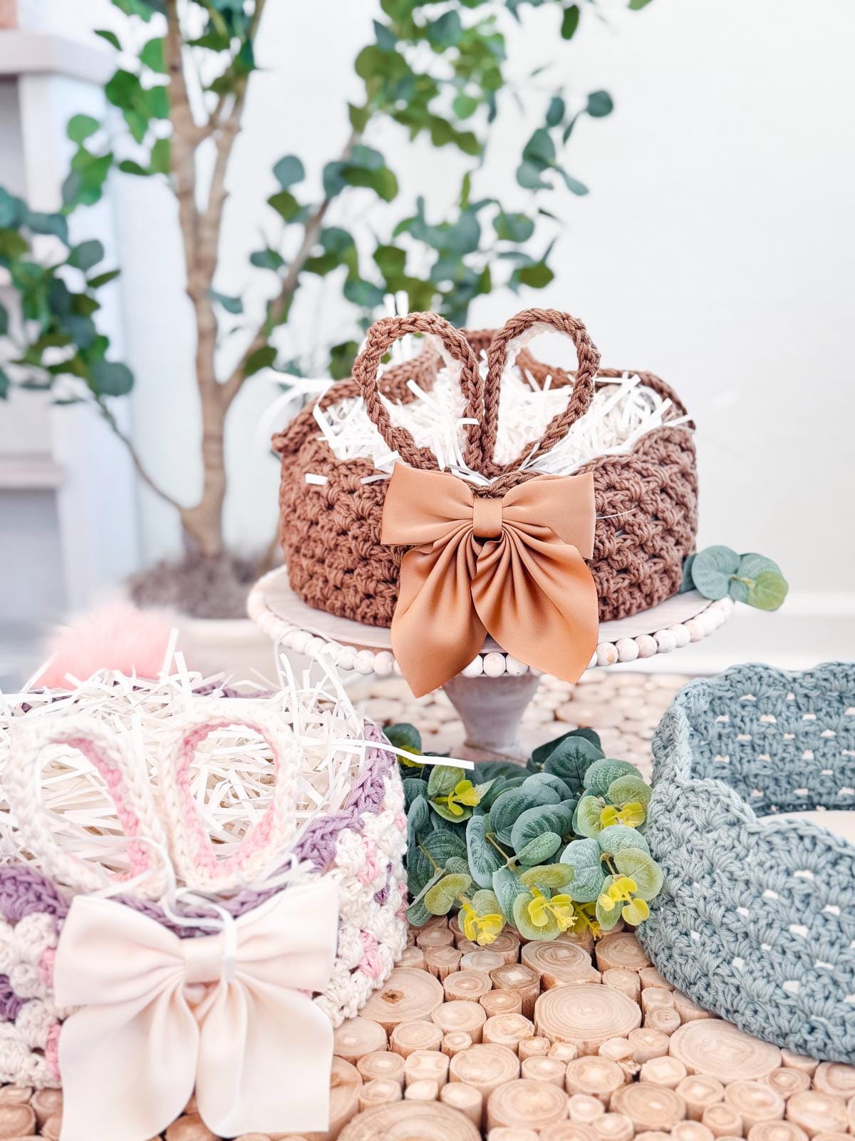 Crocheted baskets shaped like bunny ears, featuring pink and brown bows, sit on a wooden surface with decorative greenery and white shredded paper inside, making a charming handmade gift.