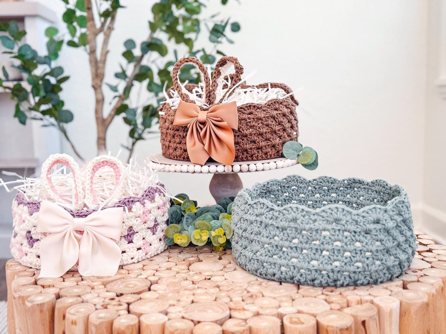 Three woven baskets with bows adorn a wooden surface, two resting on the table and one elevated on a cake stand. Greenery decorates the scene, with a tree in the background. These handmade gifts create a charming tableau perfect for any occasion.