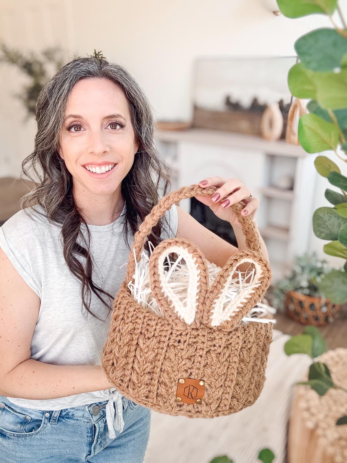 A woman with long hair holds a crocheted basket with bunny ear details, standing in a bright room adorned with plants and vintage clothes on the shelf in the background.