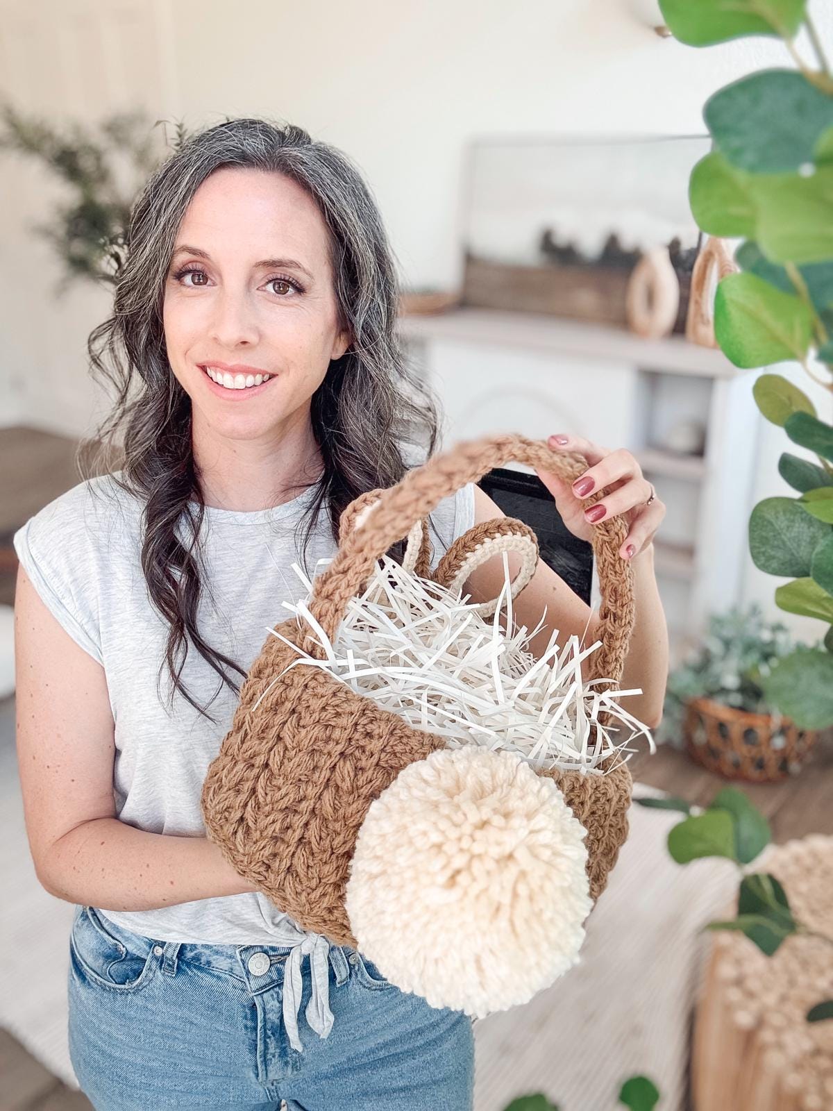 A person holds a handmade crochet easter basket with a large pom-pom, filled with shredded paper, in a softly decorated room.
