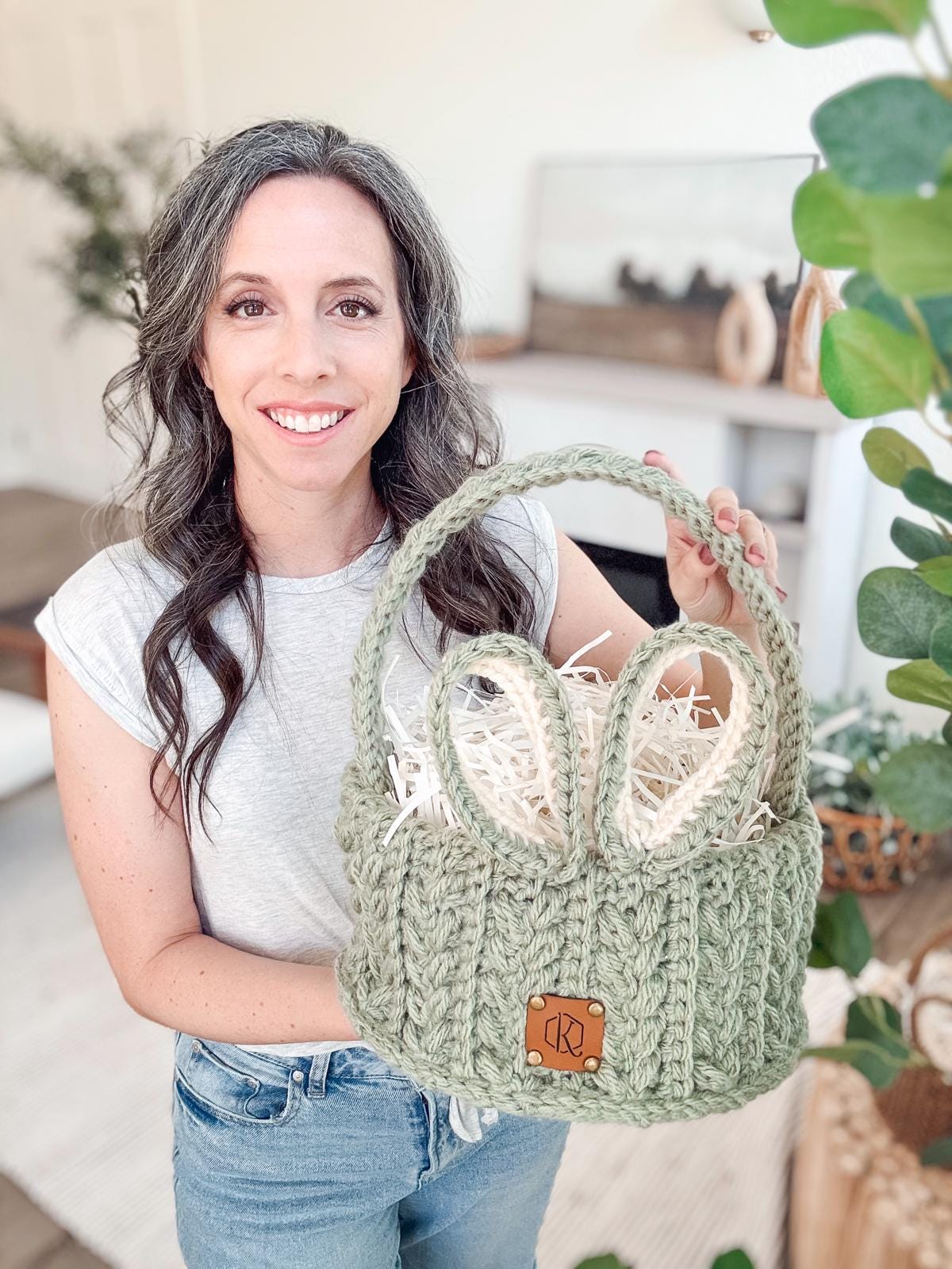 A woman, dressed in jeans and a grey tshirt, holds a crocheted green basket with bunny ears, filled with shredded paper. She stands in a room adorned with plants.