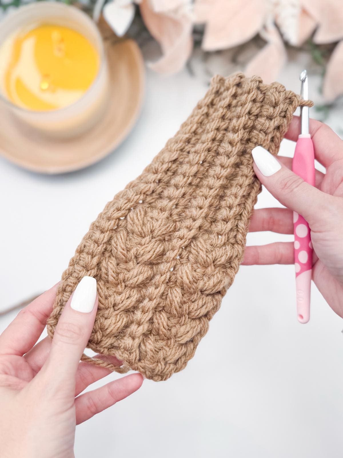 Hands delicately grip a crochet project with tan yarn, paired with a pink-handled crochet hook and the warm glow of a lit candle. This scene offers inspiration for crafting the perfect handmade gift.