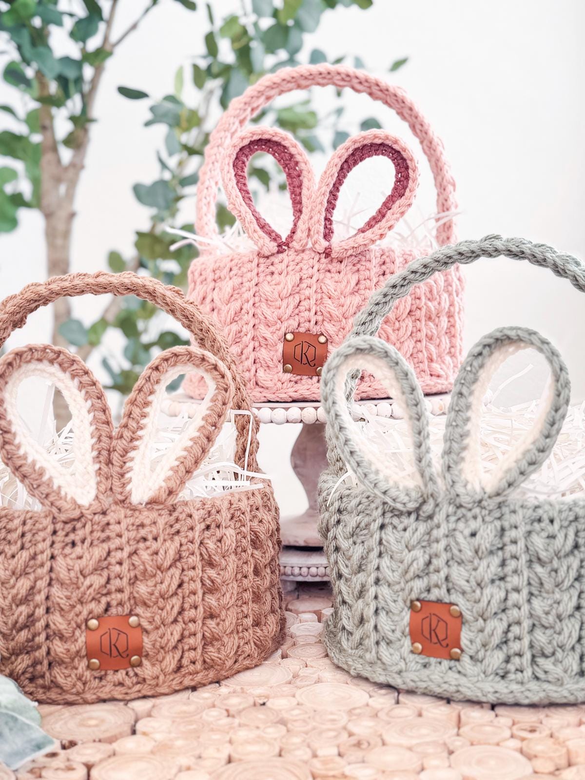 Three crochet baskets with bunny ear designs in pink, brown, and gray are displayed on a wooden surface with trees in the background. These handmade easter basket gifts add charm and whimsy.