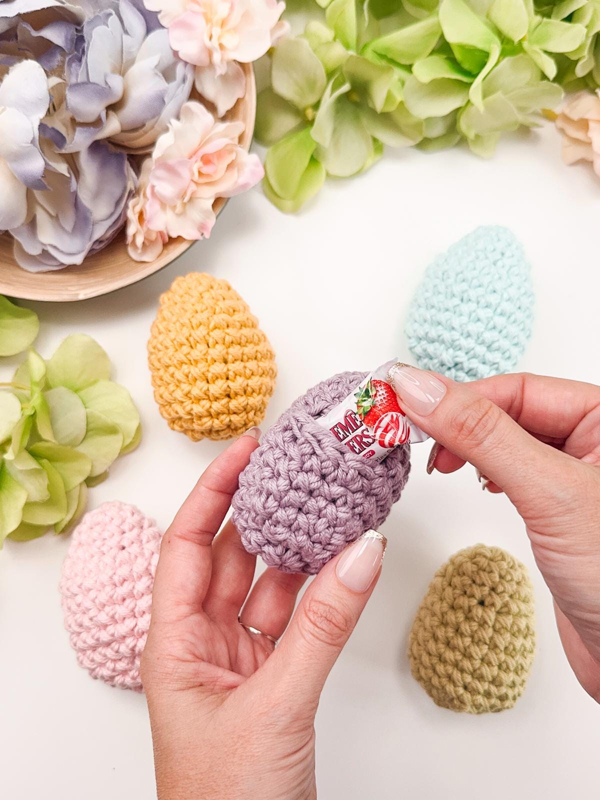 Hands opening a crocheted egg with a candy inside, surrounded by other colorful crocheted eggs and flowers—a delightful handmade gift perfect for any occasion.
